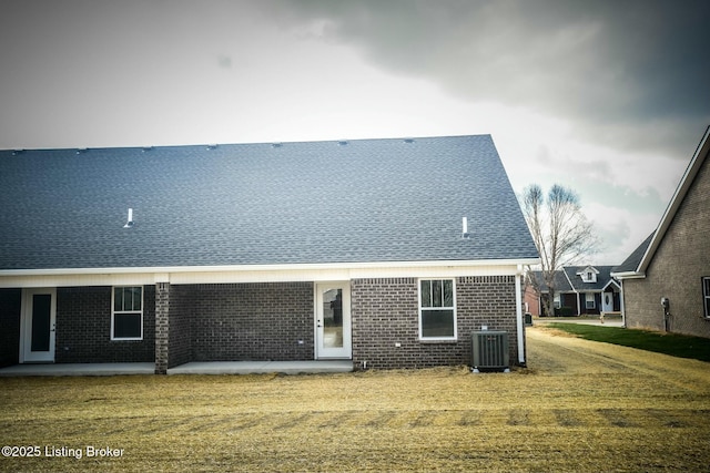 back of house with a lawn, central AC, a shingled roof, brick siding, and a patio area