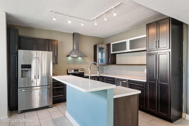kitchen featuring light countertops, stainless steel appliances, a center island with sink, glass insert cabinets, and wall chimney range hood