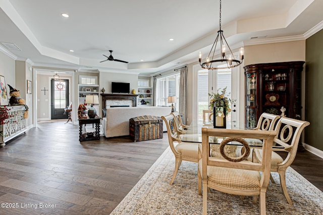dining room with ceiling fan with notable chandelier, a fireplace, a tray ceiling, visible vents, and wood finished floors