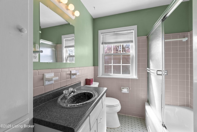 bathroom with combined bath / shower with glass door, toilet, tile walls, vanity, and a wainscoted wall