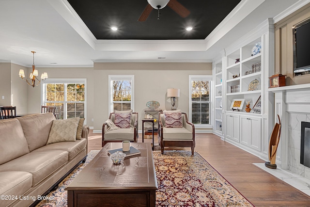 living area with crown molding, a raised ceiling, a fireplace, wood finished floors, and ceiling fan with notable chandelier