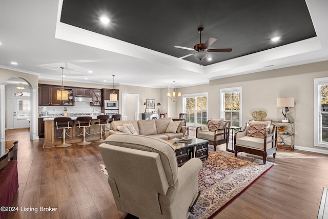 living area featuring a raised ceiling, dark wood-style flooring, ceiling fan with notable chandelier, and crown molding