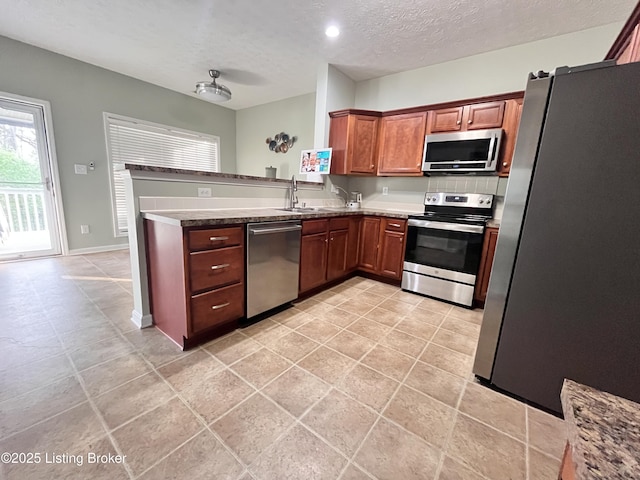 kitchen featuring stainless steel appliances, a peninsula, a sink, a textured ceiling, and dark stone countertops