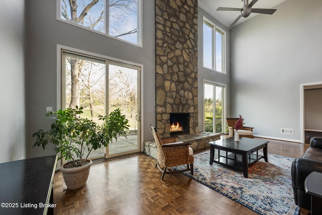 living room featuring a towering ceiling, a ceiling fan, a stone fireplace, visible vents, and baseboards