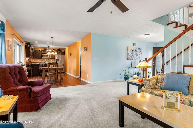 living area with ceiling fan with notable chandelier, a textured ceiling, carpet flooring, stairs, and baseboards
