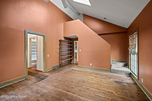 unfurnished living room with high vaulted ceiling, stairway, a skylight, wood finished floors, and baseboards