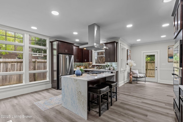 kitchen with light countertops, freestanding refrigerator, dark brown cabinets, decorative backsplash, and island range hood