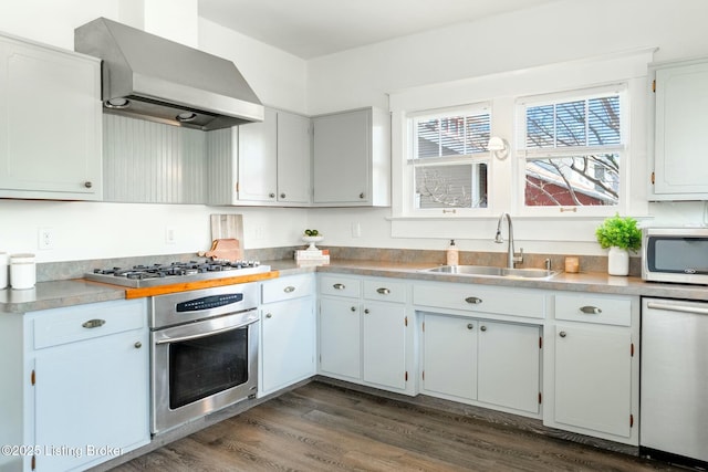 kitchen with wall chimney exhaust hood, a sink, light countertops, appliances with stainless steel finishes, and dark wood-type flooring