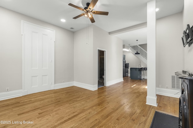 unfurnished living room featuring stairway, light wood-style flooring, baseboards, and ceiling fan