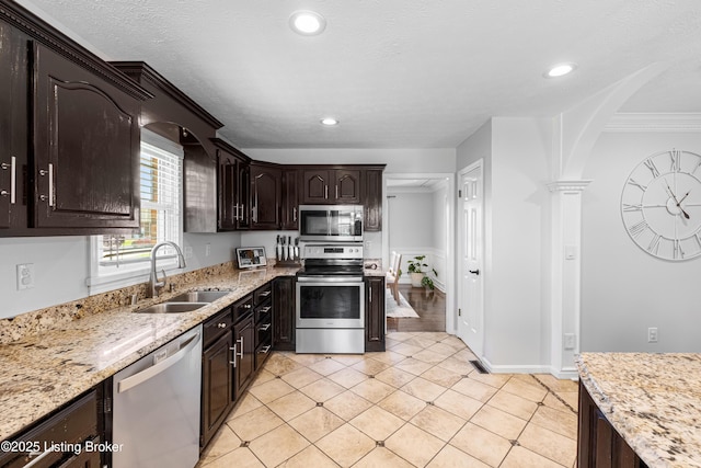 kitchen featuring a sink, appliances with stainless steel finishes, decorative columns, dark brown cabinets, and light stone counters