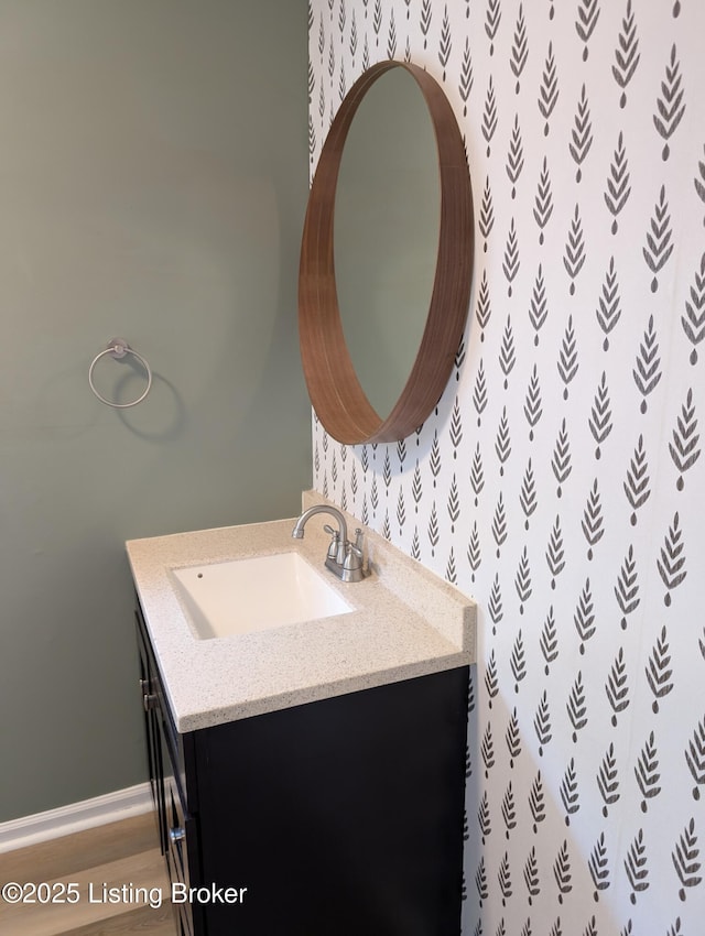 bathroom featuring vanity, baseboards, wallpapered walls, and wood finished floors