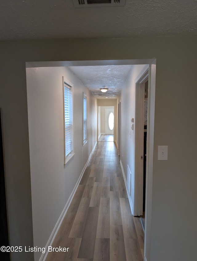 hallway with a textured ceiling, wood finished floors, baseboards, and visible vents