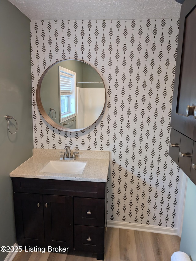 full bath with a textured ceiling, wallpapered walls, vanity, and wood finished floors
