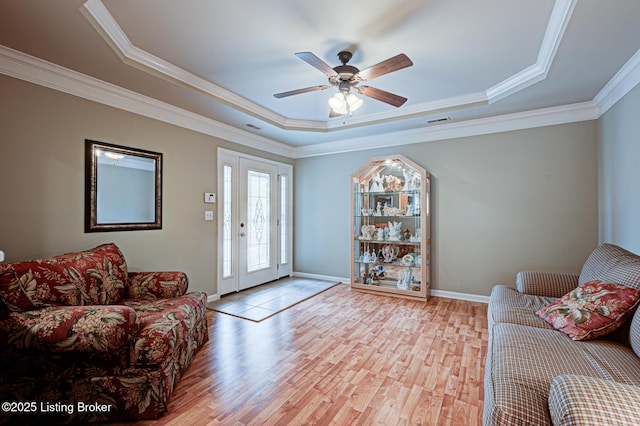 living room with baseboards, a raised ceiling, crown molding, light wood-style floors, and ceiling fan