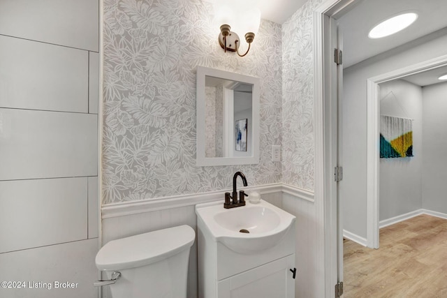 half bathroom with wood finished floors, wallpapered walls, vanity, a wainscoted wall, and toilet