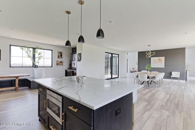 kitchen featuring light wood finished floors, stainless steel microwave, dark cabinets, a kitchen island, and hanging light fixtures