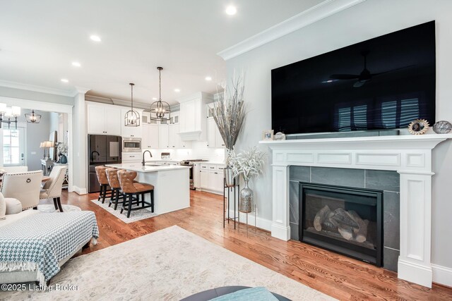 living area with ceiling fan with notable chandelier, a glass covered fireplace, light wood-style flooring, ornamental molding, and recessed lighting