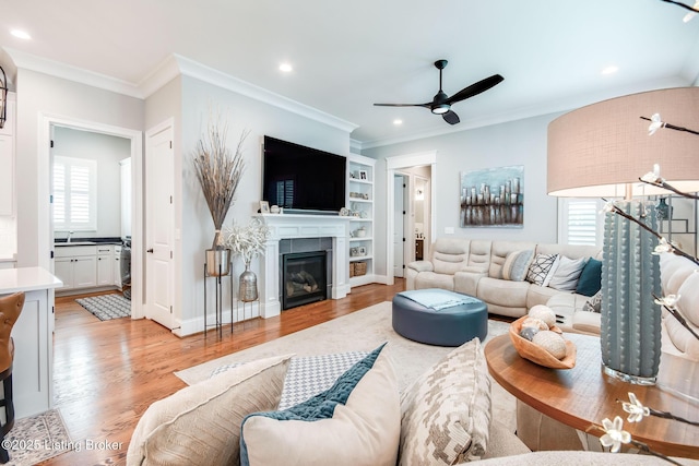 living room with light wood-type flooring, a glass covered fireplace, recessed lighting, ceiling fan, and crown molding