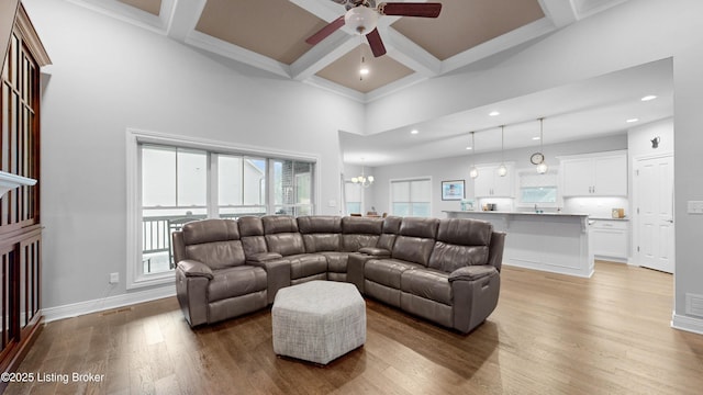 living area with coffered ceiling, beamed ceiling, ceiling fan with notable chandelier, light wood finished floors, and a high ceiling
