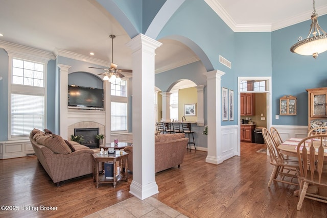 interior space with a ceiling fan, wood finished floors, a tiled fireplace, visible vents, and decorative columns