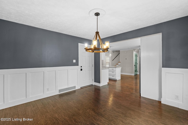 unfurnished dining area featuring a wainscoted wall, dark wood finished floors, visible vents, an inviting chandelier, and stairs
