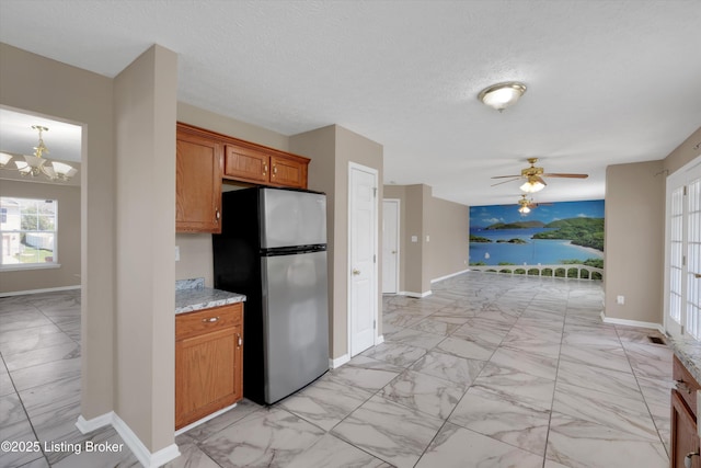 kitchen with marble finish floor, freestanding refrigerator, baseboards, brown cabinetry, and ceiling fan with notable chandelier