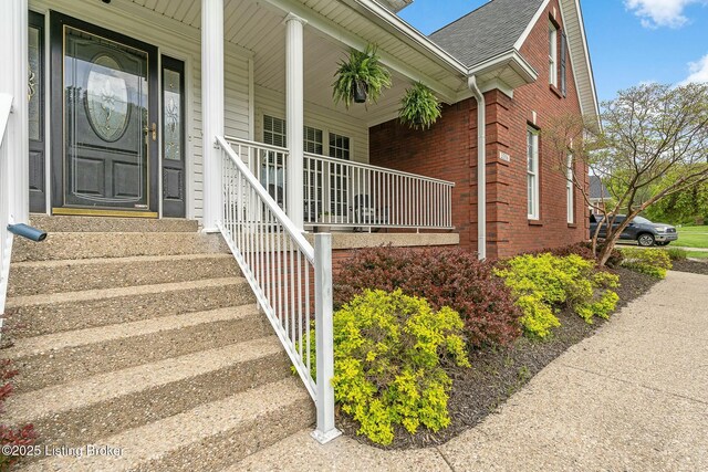 entrance to property featuring a porch, a shingled roof, and brick siding