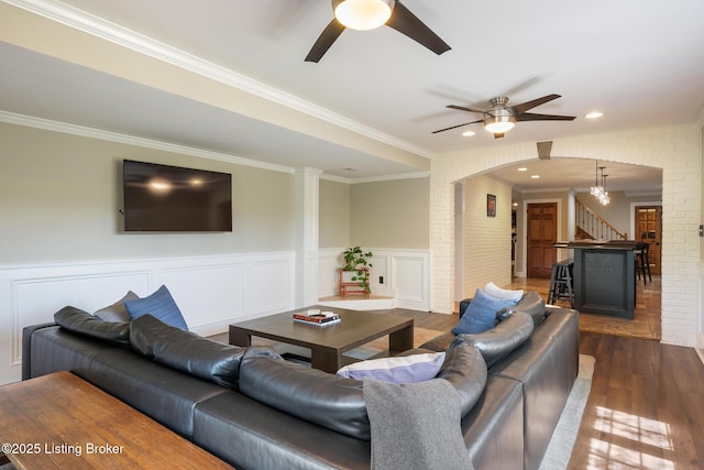 living room featuring a wainscoted wall, arched walkways, ornamental molding, dark wood finished floors, and ceiling fan with notable chandelier