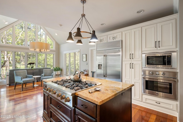 kitchen with light stone counters, built in appliances, hardwood / wood-style floors, dark brown cabinetry, and a notable chandelier