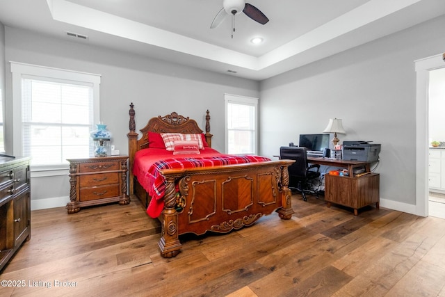 bedroom featuring a tray ceiling, wood finished floors, visible vents, baseboards, and ceiling fan