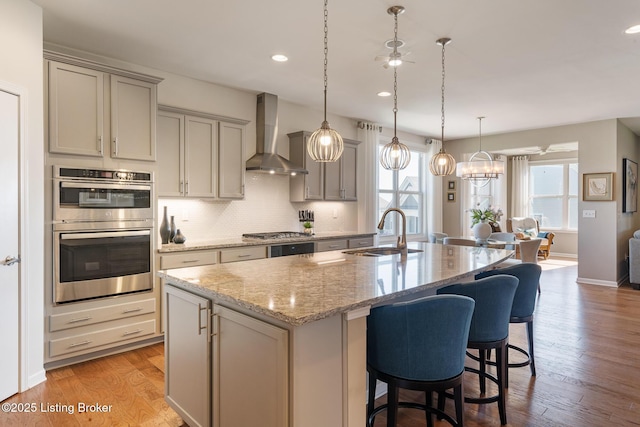 kitchen featuring wall chimney range hood, appliances with stainless steel finishes, a sink, gray cabinets, and a center island with sink