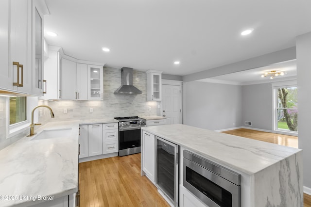 kitchen with wall chimney range hood, a sink, light wood-style flooring, appliances with stainless steel finishes, and wine cooler