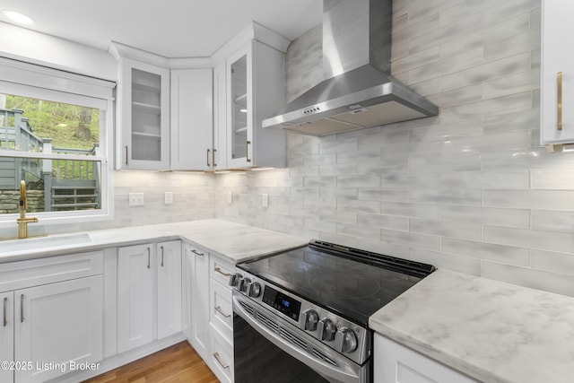 kitchen with wall chimney range hood, a sink, stainless steel electric range, backsplash, and white cabinetry