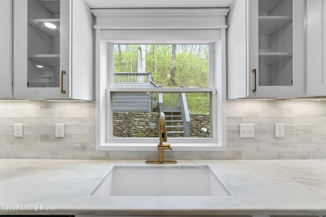interior details with light stone counters, tasteful backsplash, a sink, glass insert cabinets, and white cabinetry