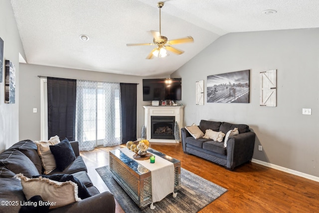 living area featuring wood finished floors, ceiling fan, a fireplace with raised hearth, baseboards, and lofted ceiling