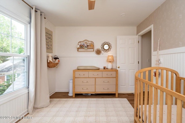 bedroom with wood finished floors, a wainscoted wall, a ceiling fan, and a crib