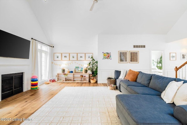 living room with wood finished floors, a wainscoted wall, a fireplace, visible vents, and high vaulted ceiling