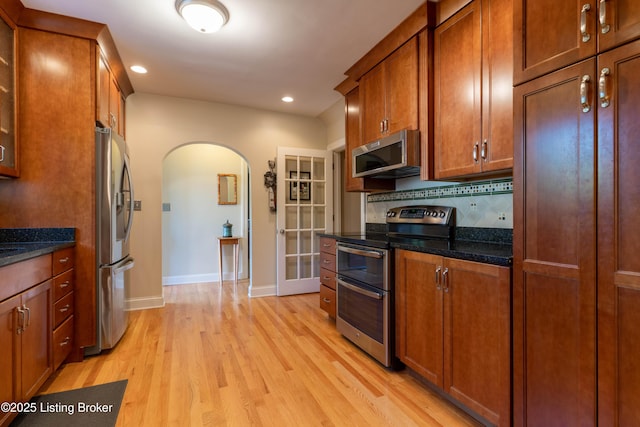 kitchen with arched walkways, brown cabinetry, appliances with stainless steel finishes, light wood finished floors, and baseboards