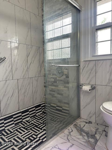 bathroom with marble finish floor, toilet, a stall shower, and tile walls