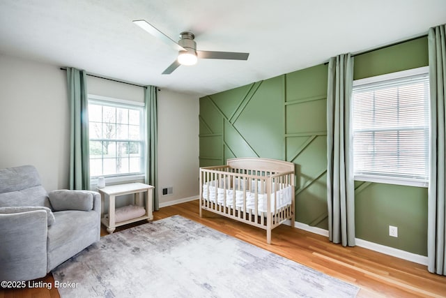 bedroom featuring wood finished floors, a nursery area, baseboards, visible vents, and a ceiling fan