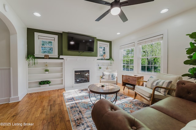 living room with a glass covered fireplace, wood finished floors, ceiling fan, recessed lighting, and arched walkways
