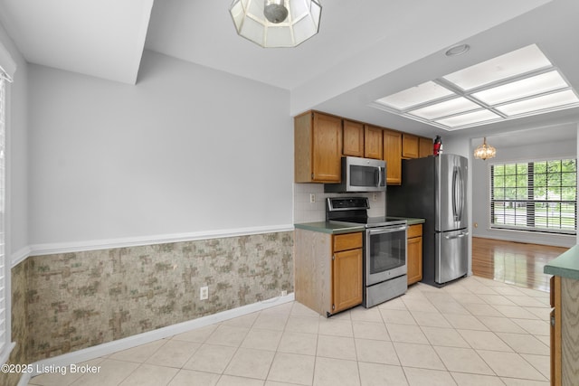 kitchen with brown cabinetry, appliances with stainless steel finishes, baseboards, an inviting chandelier, and wallpapered walls