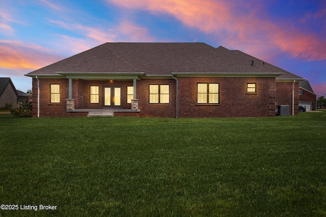 back of property at dusk with a lawn, covered porch, cooling unit, and roof with shingles