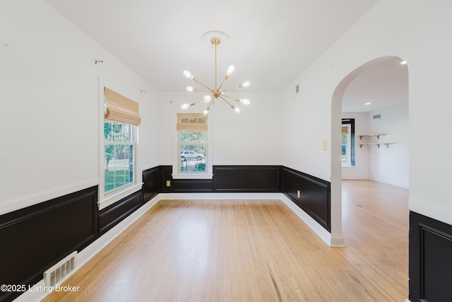 unfurnished dining area featuring light wood-style flooring, visible vents, wainscoting, arched walkways, and a chandelier