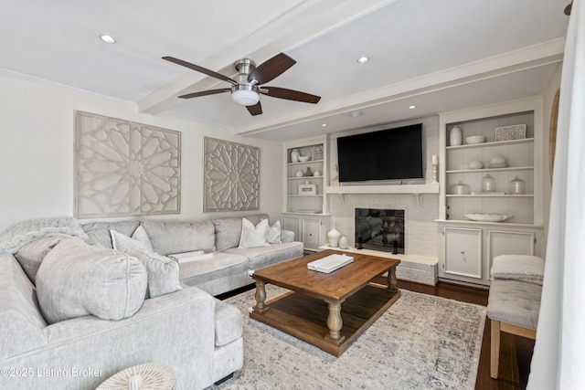living room featuring crown molding, beam ceiling, a brick fireplace, built in shelves, and a ceiling fan