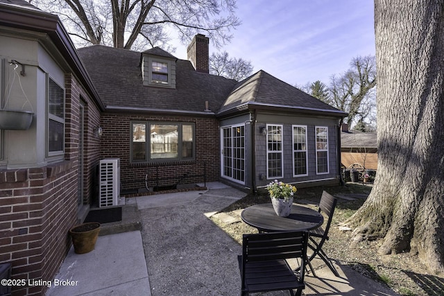 rear view of house featuring brick siding, a patio area, roof with shingles, and a chimney
