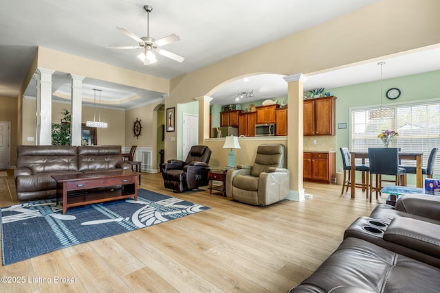 living area with arched walkways, light wood-style flooring, ceiling fan with notable chandelier, ornate columns, and a tray ceiling