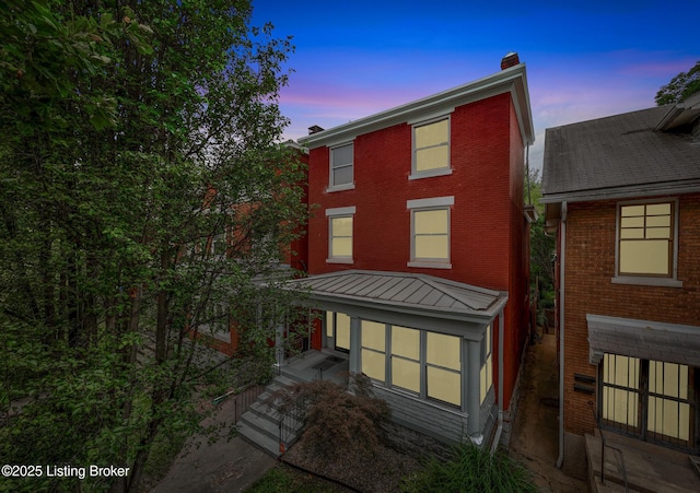 back of property at dusk featuring a chimney, brick siding, and a standing seam roof