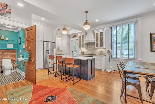 kitchen featuring stainless steel refrigerator with ice dispenser, custom range hood, light wood-type flooring, a sink, and white cabinets
