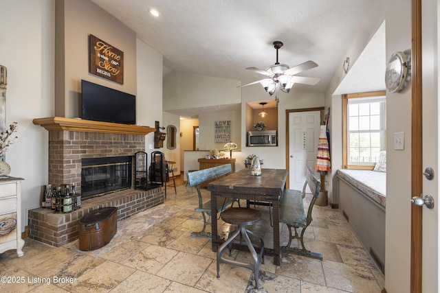 dining area with ceiling fan, a fireplace, a textured ceiling, stone tile floors, and lofted ceiling